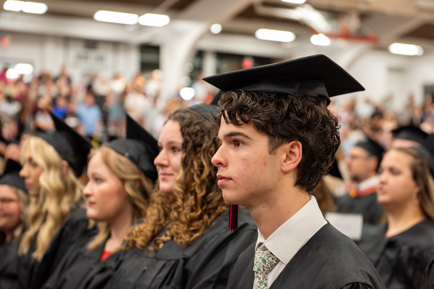 A side-profile shot of a row of graduates seated during an indoor commencement ceremony. In the foreground, a young male graduate with curly dark hair looks forward attentively, wearing a black graduation cap, gown, white dress shirt, and a light-patterned tie. Next to him sits a female graduate with curly hair, followed by other seated graduates extending to the left. In the blurred background, a large crowd of spectators fills the stands of a gym or auditorium under bright indoor lighting.