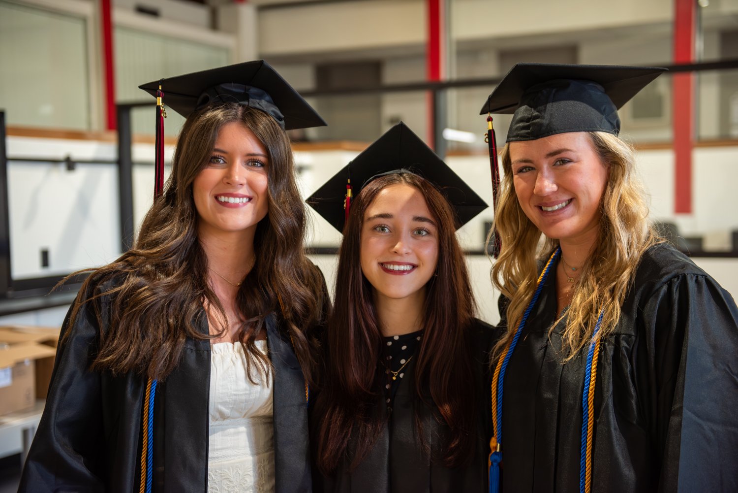A close-up portrait of three smiling female graduates standing side-by-side indoors. All three are wearing black graduation caps and gowns. The graduate on the left has long, wavy brown hair and wears a white dress under her gown. The graduate in the center has straight dark hair and a darker complexion, wearing a black polka-dot top. The graduate on the right has blonde hair and wears blue and gold honor cords draped over her shoulders. The background shows a modern indoor facility with white walls, glass partitions, and red structural pillars.