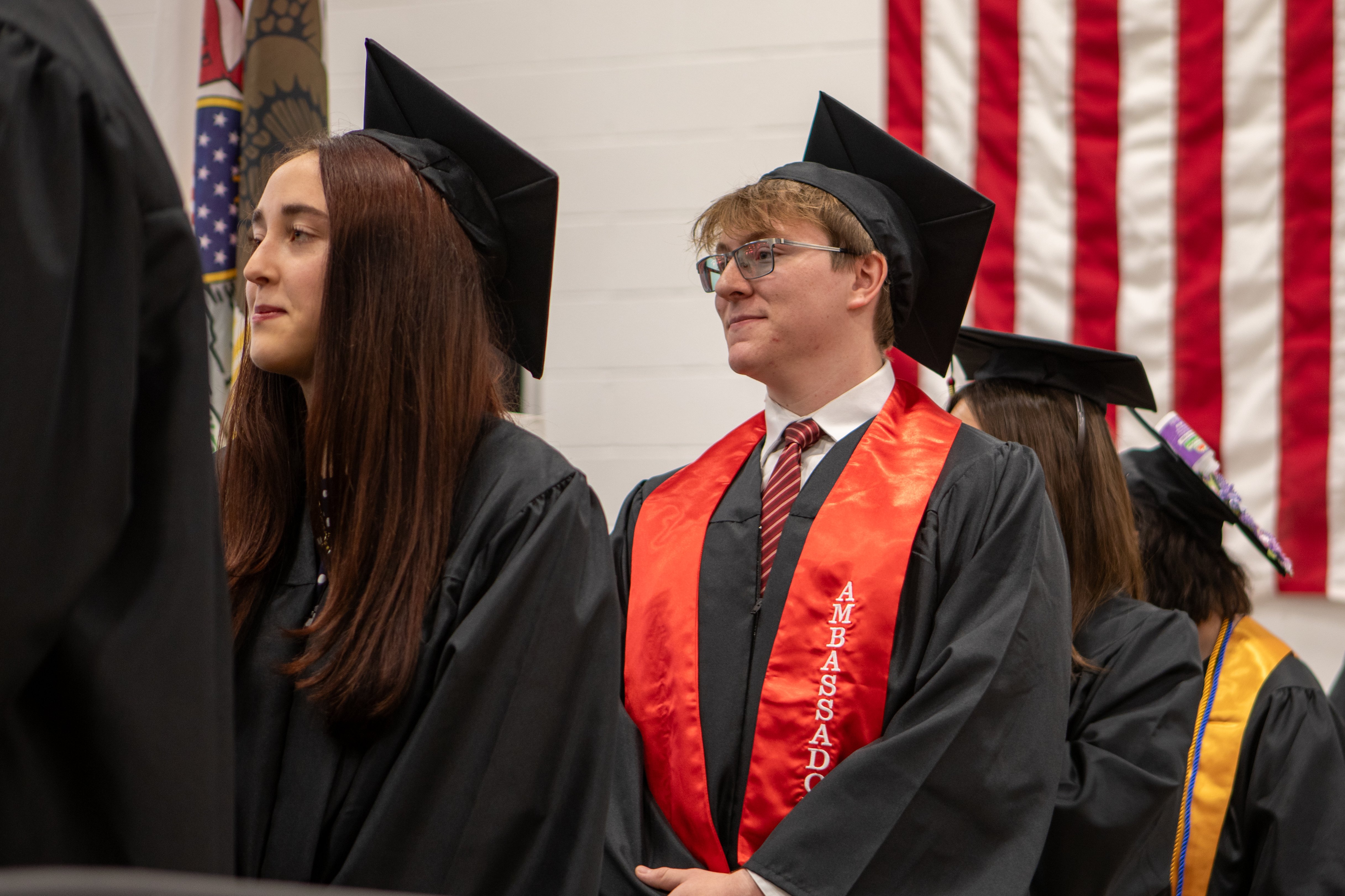 A line of graduates standing indoors during a commencement ceremony, all wearing black academic caps and gowns. On the left, a female graduate with straight brown hair looks forward. Next to her, a male graduate wearing glasses, a white dress shirt, and a red patterned tie smiles while wearing a bright red graduation stole with white text that reads "AMBASSADOR". An American flag hangs on the wall in the background to the right.