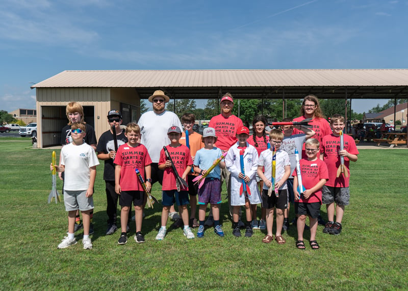 A large group of children and adult instructors stand together outdoors on a sunny lawn in front of a pavilion. Several kids wear matching red "Summer Starts at The Lake" t-shirts, and each child holds up a colorful toy model rocket they built. One child near the center wears a white lab coat.