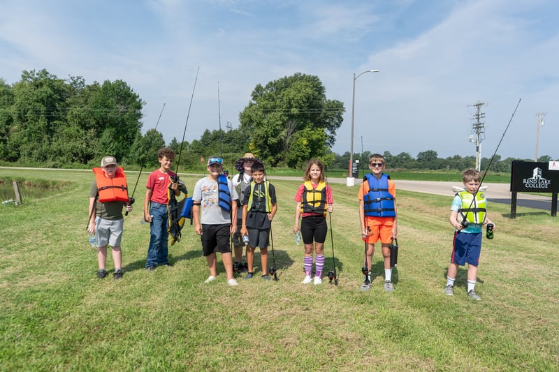 A group of young kids holding golf clubs poses on a green course with three instructors standing behind them on a sunny day.
