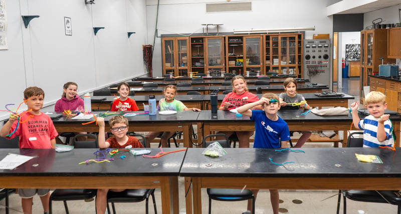 A group of young students sits behind long black-topped lab tables in a science classroom, smiling and holding up bent, colorful pipe cleaners. Several kids wear red t-shirts, including one that reads "Summer Starts at The Lake." Wooden cabinets and laboratory equipment line the back wall of the room.