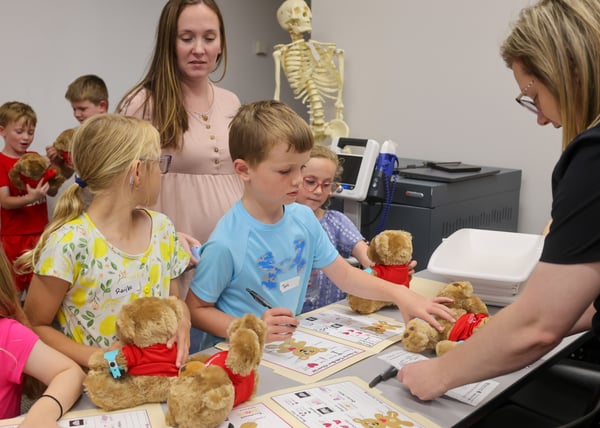 Children gather around a table with Teddy Bear Clinic check-in worksheets, writing on paper forms while examining plush teddy bears wearing mini hospital ID bands and red shirts. An instructor stands nearby assisting them, with a full-sized human skeleton model standing in the background.