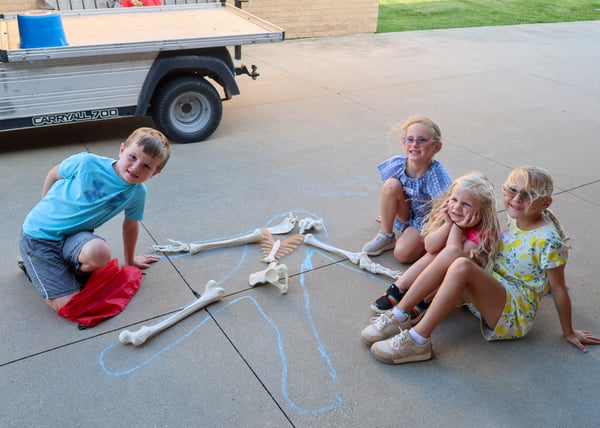 Campers sit outside around a sidewalk chalk outline filled in with plastic model bones during an anatomy activity.