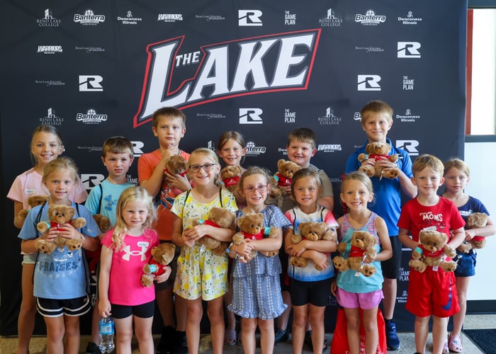 A group photo of about twelve children standing together in front of a dark backdrop featuring "The Lake" and Rend Lake College logos. Each camper proudly holds a brown plush teddy bear wearing a red shirt and a blue hospital wristband.