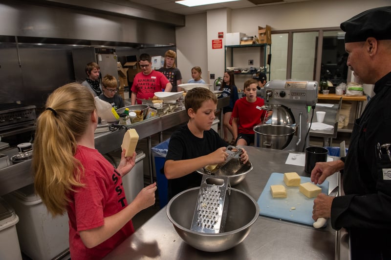 A chef in black uniform instructs a group of young students in a commercial kitchen as they grate block cheese into stainless steel bowls.
