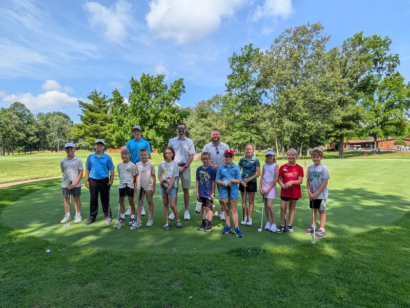 A group of young kids holding golf clubs poses on a green course with three instructors standing behind them on a sunny day.