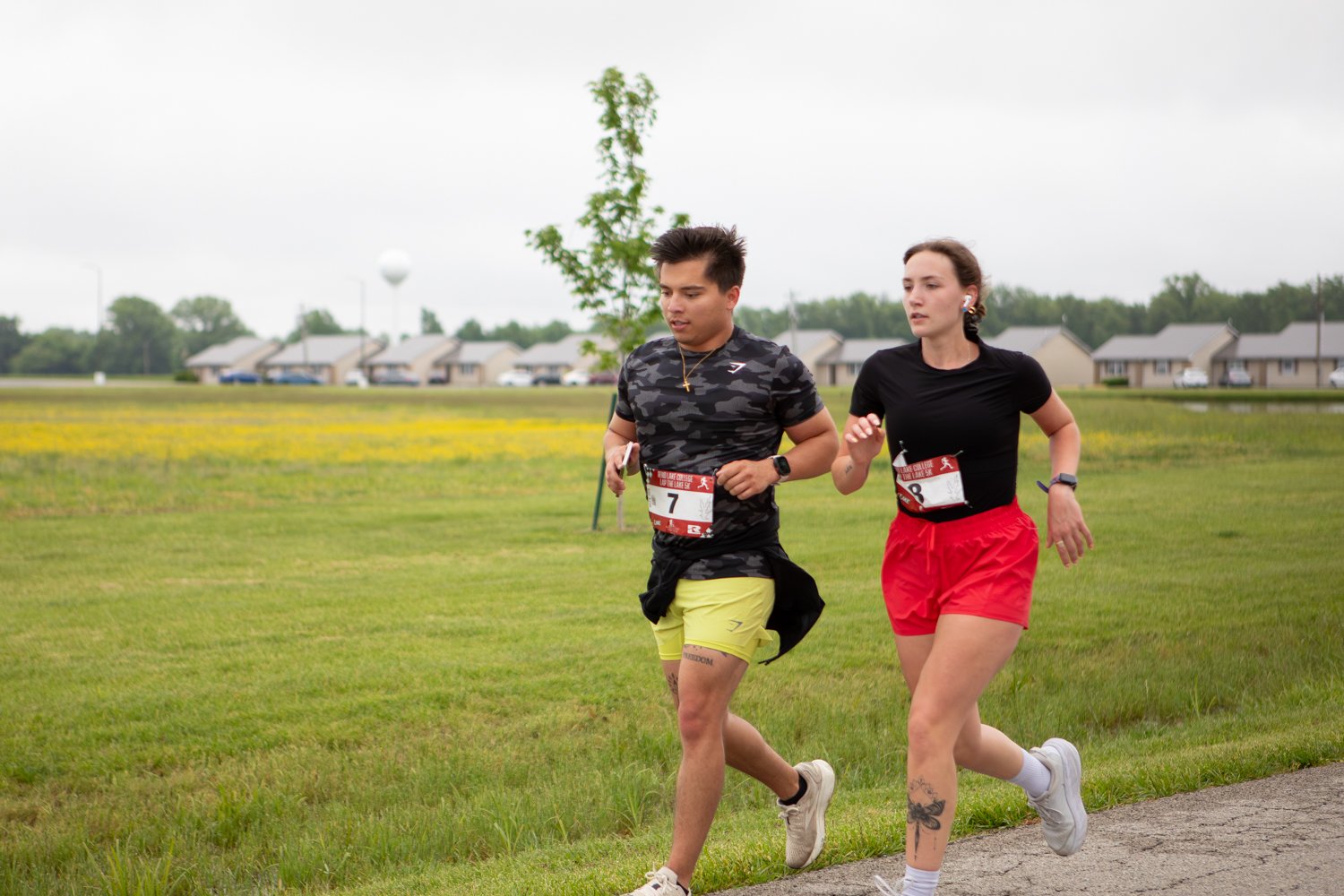A young man wearing a camouflage t-shirt and yellow shorts with race bib number 7 runs side-by-side with a young woman wearing a black t-shirt and red shorts along a campus path. A row of single-story campus housing units is visible in the background across an open green field.