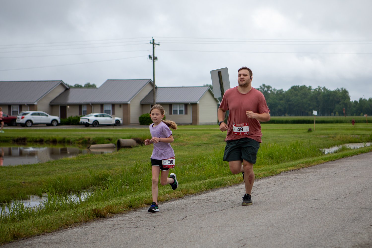 A young girl wearing race bib number 30 runs down an asphalt campus road ahead of a man in a pink t-shirt and dark shorts wearing race bib number 49. In the background, modern campus housing complexes sit next to a small pond under a grey sky.