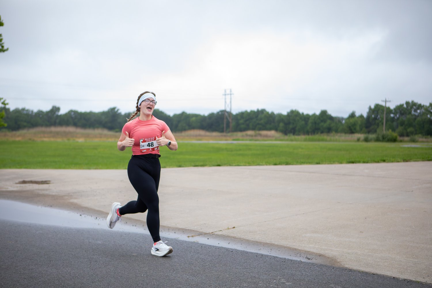 A female runner wearing race bib number 48, a pink t-shirt, black leggings, and a white headband smiles widely and gives a double thumbs-up while running along a paved path next to a wide, open grassy field under an overcast sky.