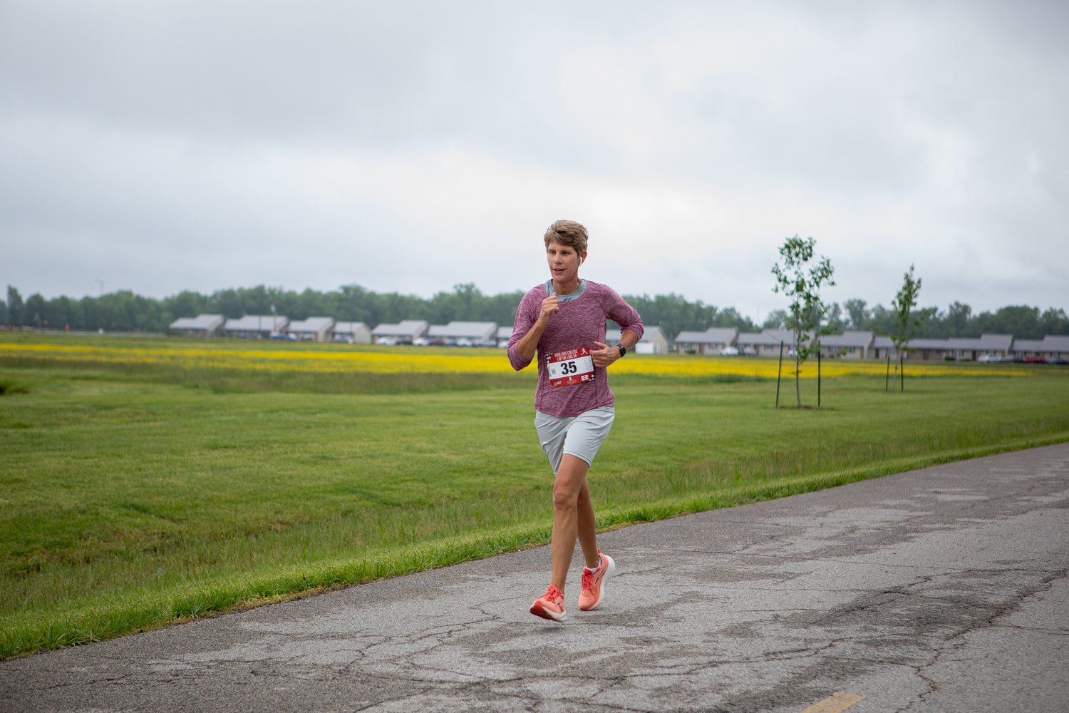 A female runner, Trish Reed, wearing race bib number 35, a heathered pink long-sleeve shirt, and grey shorts, jogs along a paved campus road. A sprawling field of green grass and yellow wildflowers sets the background, with campus housing units visible in the distance.