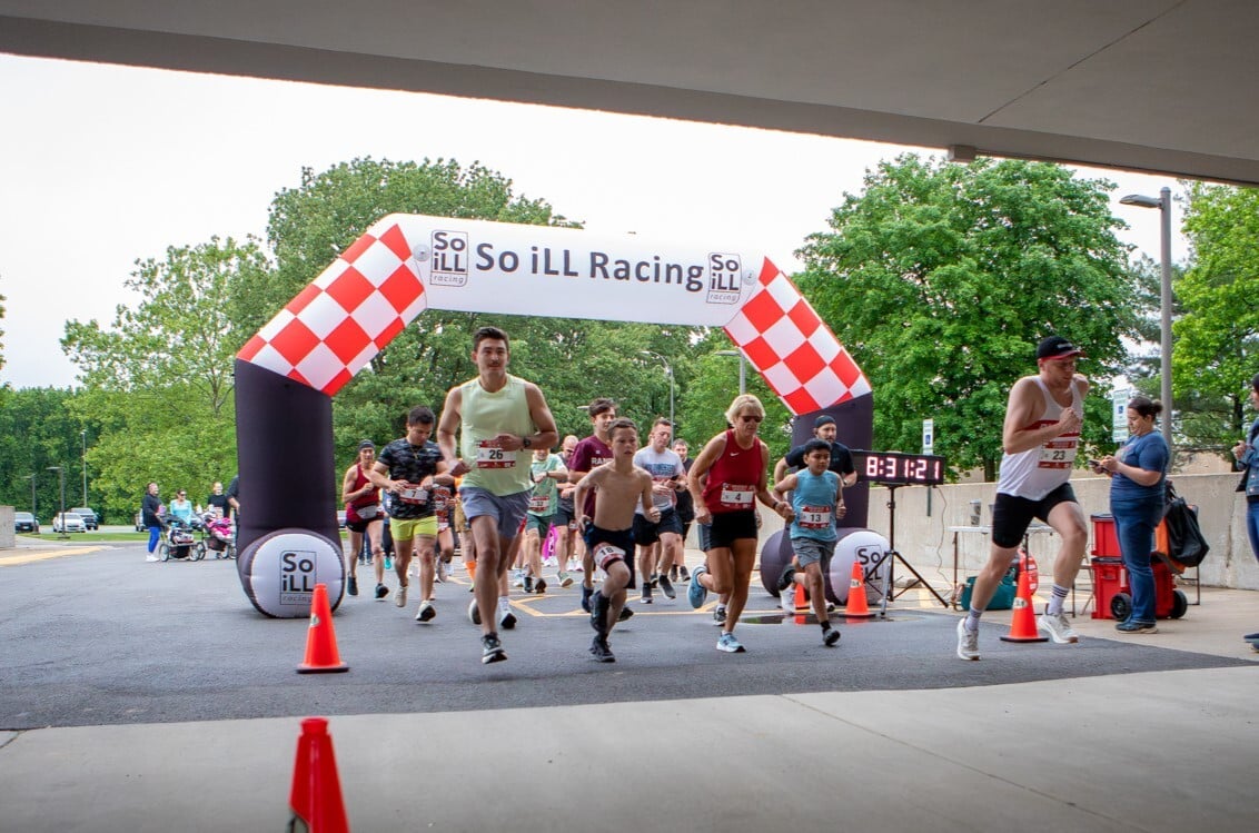 A crowd of diverse runners bursts forward from the starting line of the race, passing under a large, inflated black-and-white checkered archway that reads "So iLL Racing." Orange traffic cones mark the course boundaries, and spectators look on from the side.