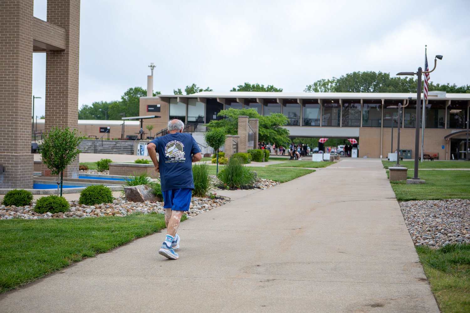 Rear view of a senior male runner wearing a dark blue t-shirt and blue shorts as he jogs down the wide concrete walkway of Warrior Way. Modern brick campus buildings, manicured green lawns, and a tall flagpole flying the American flag are visible under an overcast sky.