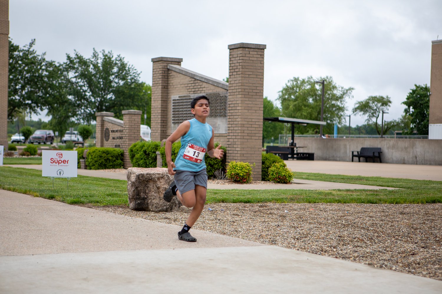 A young boy wearing a light blue sleeveless shirt, grey shorts, and race bib number 13 runs past a large brick monument on a campus walkway. A white event sign reading "The Super Patch Company" stands on the lawn to the left against a cloudy sky.