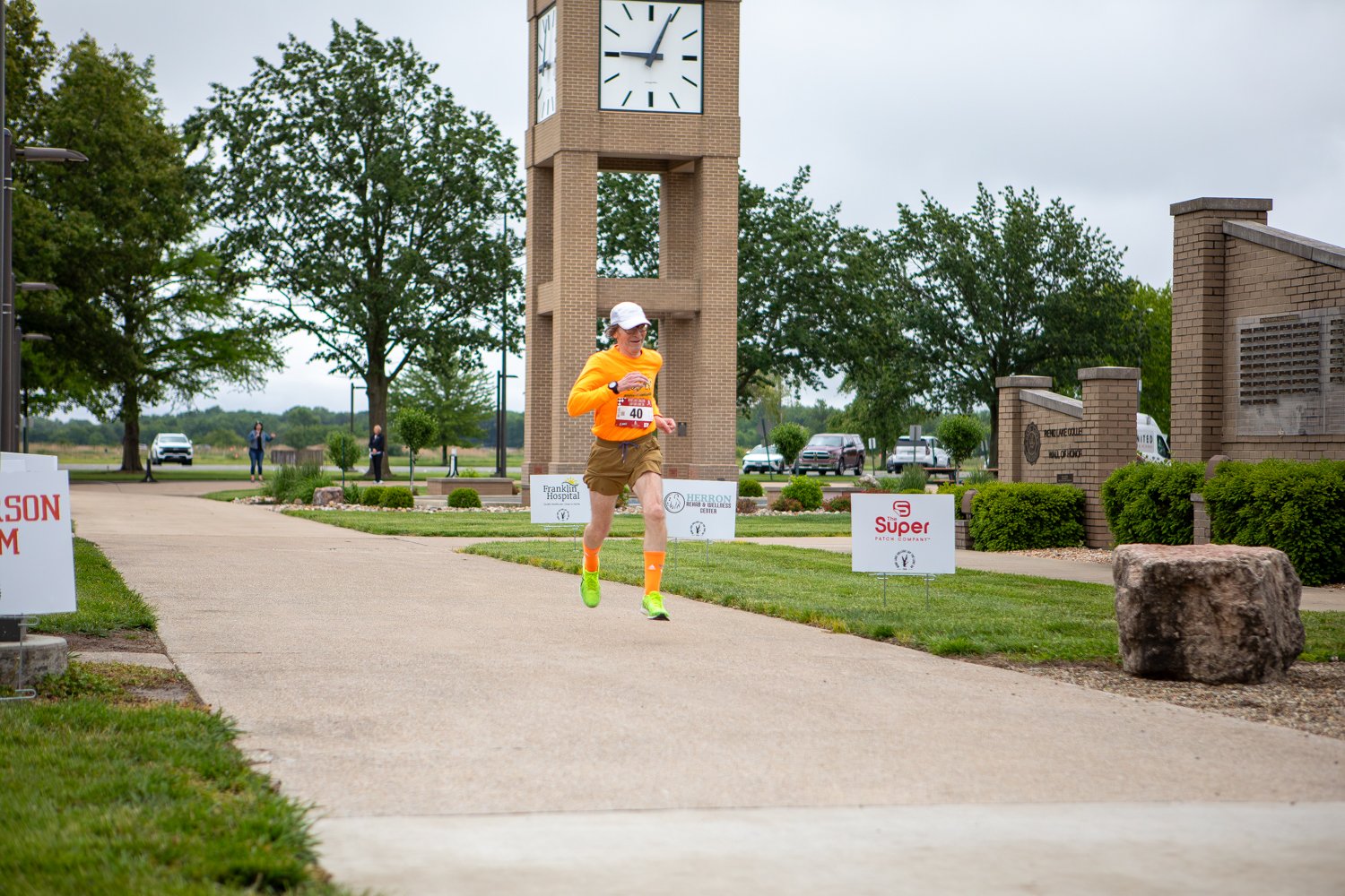 A male runner wearing an orange long-sleeve shirt, brown shorts, bright orange high socks, and a white cap jogs down Warrior Way. A tall, brick clock tower stands prominently in the background, surrounded by green trees and event sponsor signs under an overcast sky.