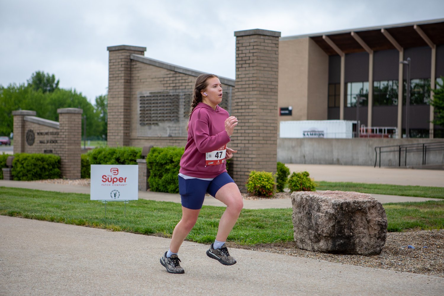 A young woman wearing a mauve hoodie, blue athletic shorts, and race bib number 47 runs along a campus path. In the background, brick architectural features, a large stone, and a modern college building are visible under a grey sky.