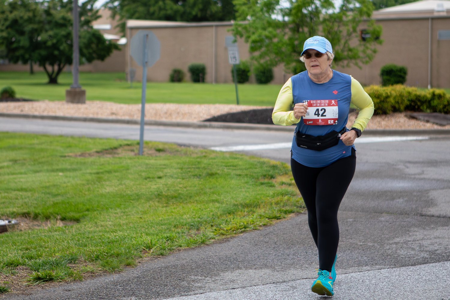 A female runner wearing race bib number 42, a blue sleeveless top over a yellow long-sleeve shirt, black leggings, and a light blue cap, running along a curved asphalt campus road. A grassy field and college buildings line the background under a cloudy sky.