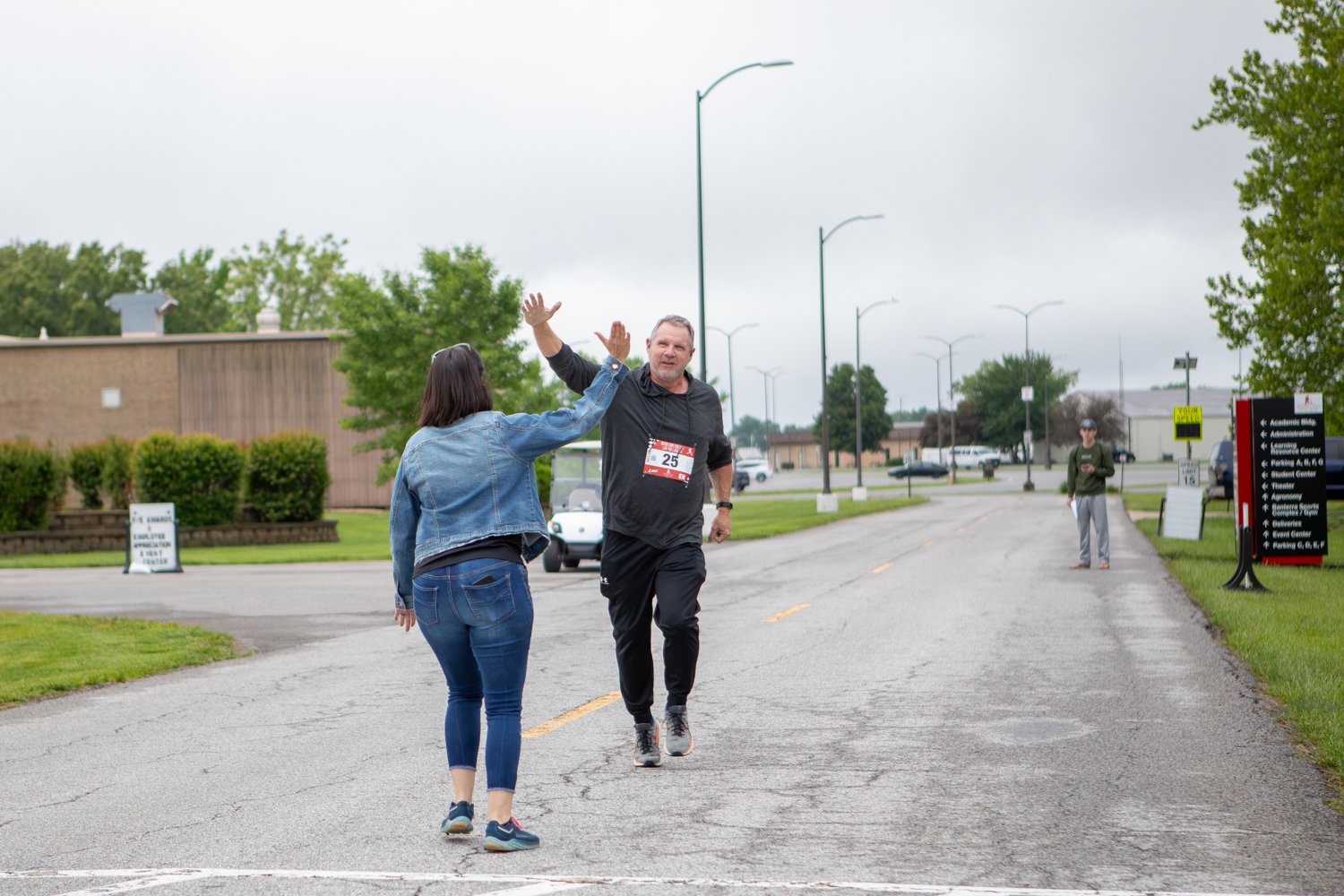 A male runner, Brad Helm, wearing race bib number 25 and dressed in a black hoodie and black sweatpants, smiles as he high-fives a woman in a denim jacket on a campus road. A campus directional sign pointing toward the Learning Resource Center and Theater stands on the right.