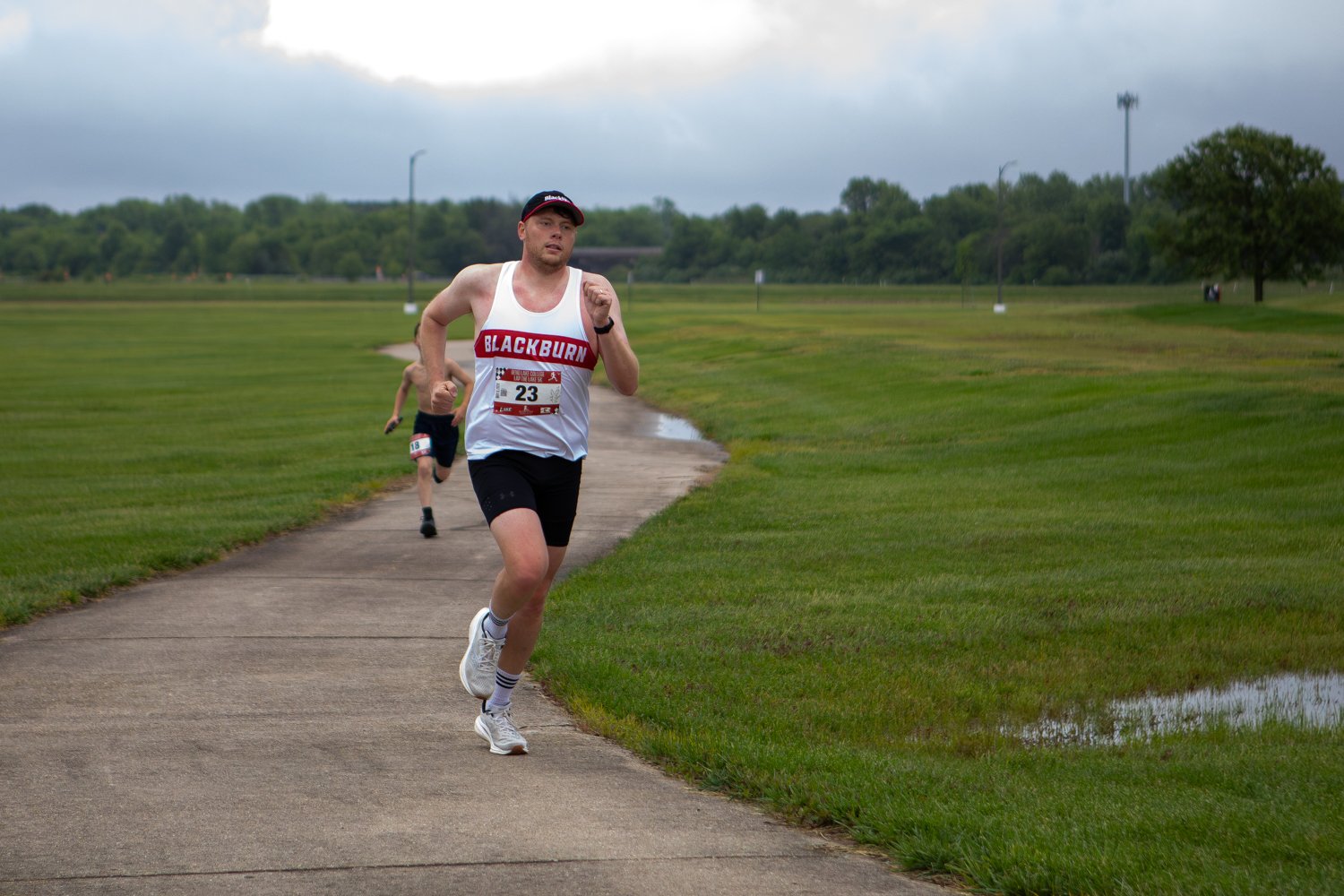 A male runner wearing a white and maroon tank top that reads "BLACKBURN" and race bib number 23 runs determinedly along a winding concrete path. A younger boy runs a short distance behind him on the path, which is bordered by expansive green grass fields under a dramatic, cloudy sky.