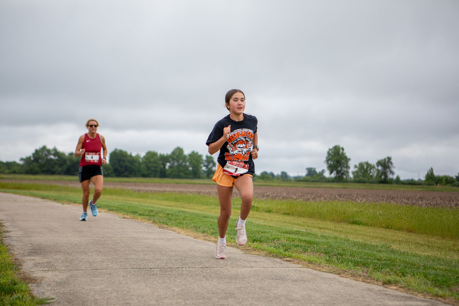 A young female runner wearing race bib number 1 and an orange-and-black graphic t-shirt leads a female runner in a maroon tank top down a paved path. The path runs alongside a wide, open green field and agricultural farmland under an overcast, cloudy sky.