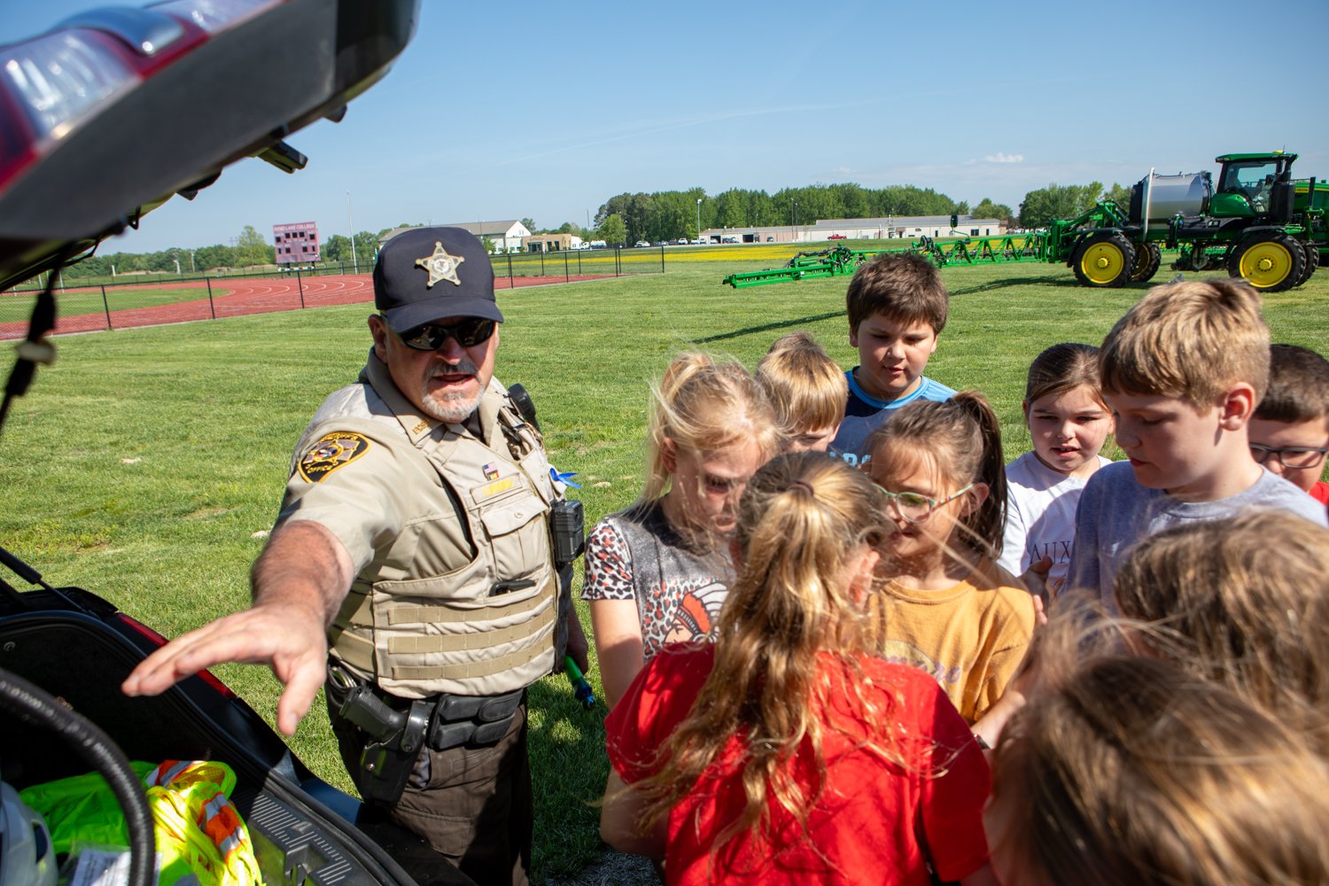  A police officer in a tan uniform and sunglasses stands outside by a car's open trunk, talking to a group of children. The kids are huddled together, looking at what is inside the trunk. They are in a grassy field with a running track and large green farm equipment in the background.