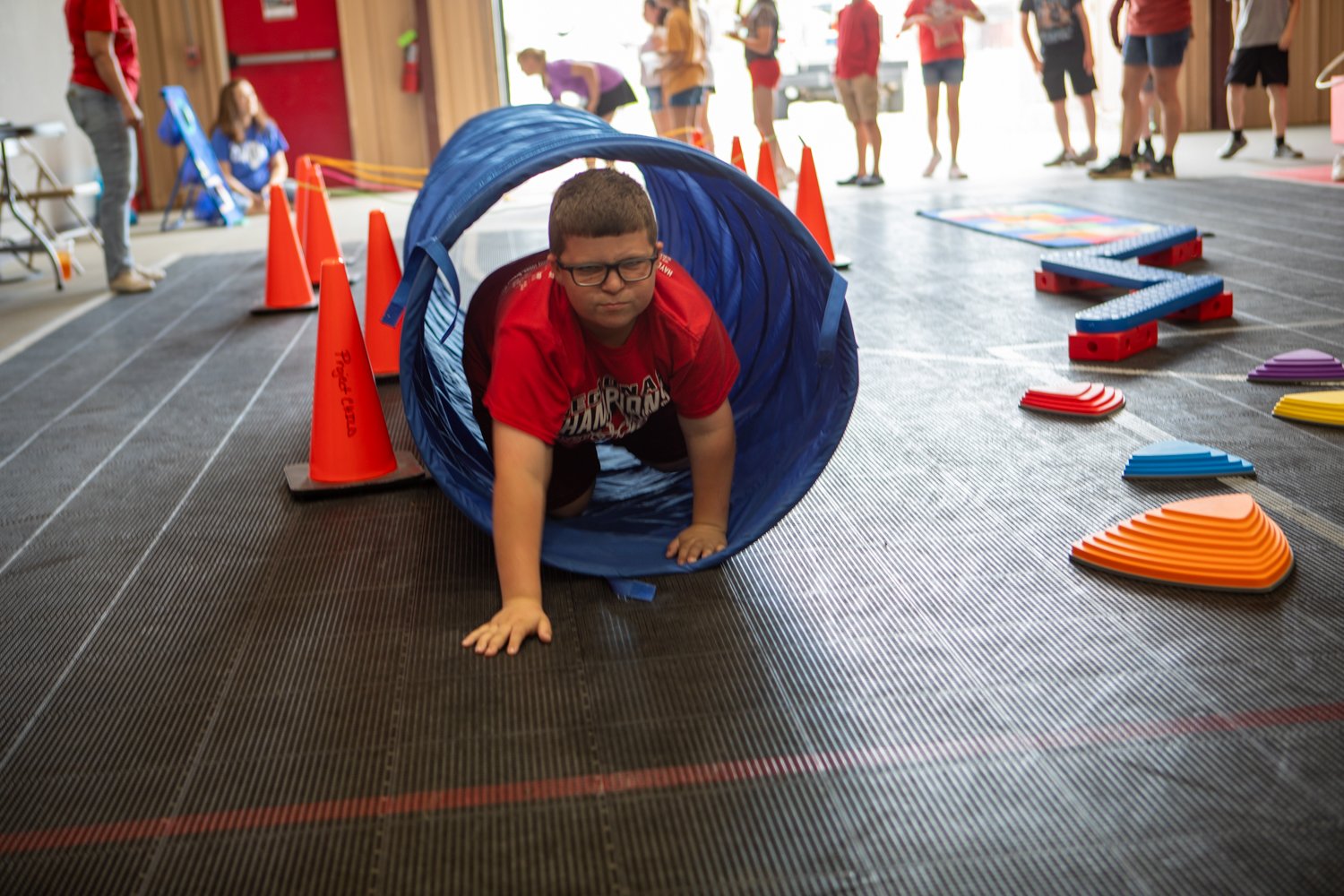 A child wearing a red shirt and glasses crawls through a blue tunnel. They are indoors on a black mat floor with other people and orange traffic cones in the background.