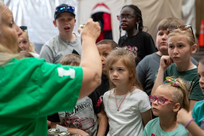 An adult in a green shirt holds up a small object for a group of curious children to examine. The kids are gathered closely, watching the demonstration with interest. On the table in front of them, several small glass jars are lined up.
