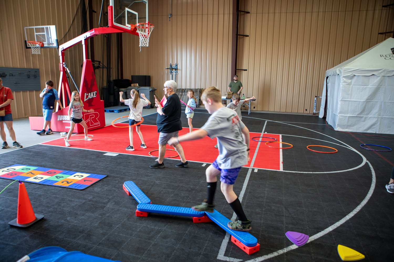 Children play on an indoor obstacle course. One boy balances on a blue beam while others play with hula hoops on a basketball court. There is an adult watching them, and a white tent is set up in the corner of the gym.