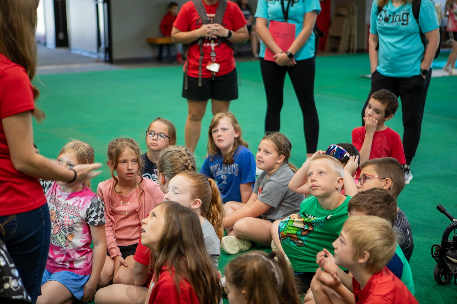 An adult in a red shirt stands on the left, speaking to a group of about 15 young children. The children are sitting on a bright green floor, looking up and listening intently.  In the background, three other adults stand and watch. The room appears to be a large indoor gym or activity center with gray walls. Everyone is dressed casually in T-shirts and shorts.