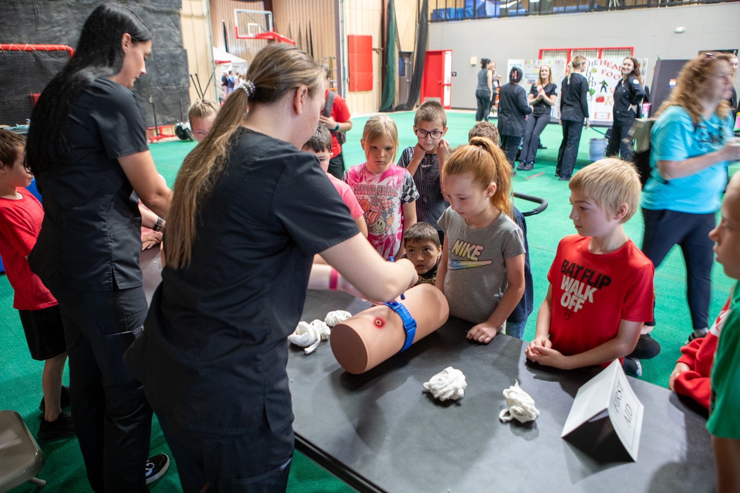 At a first aid demonstration booth, a group of children intently watches two instructors in black scrubs as they demonstrate how to use a blue tourniquet on a medical training mannequin arm. The mannequin arm features a simulated wound, and several rolls of white gauze are scattered across the black table next to a small white sign labeled "FIRST AID". The event is taking place in a large gymnasium with green flooring, where other health-related booths and participants can be seen in the background.