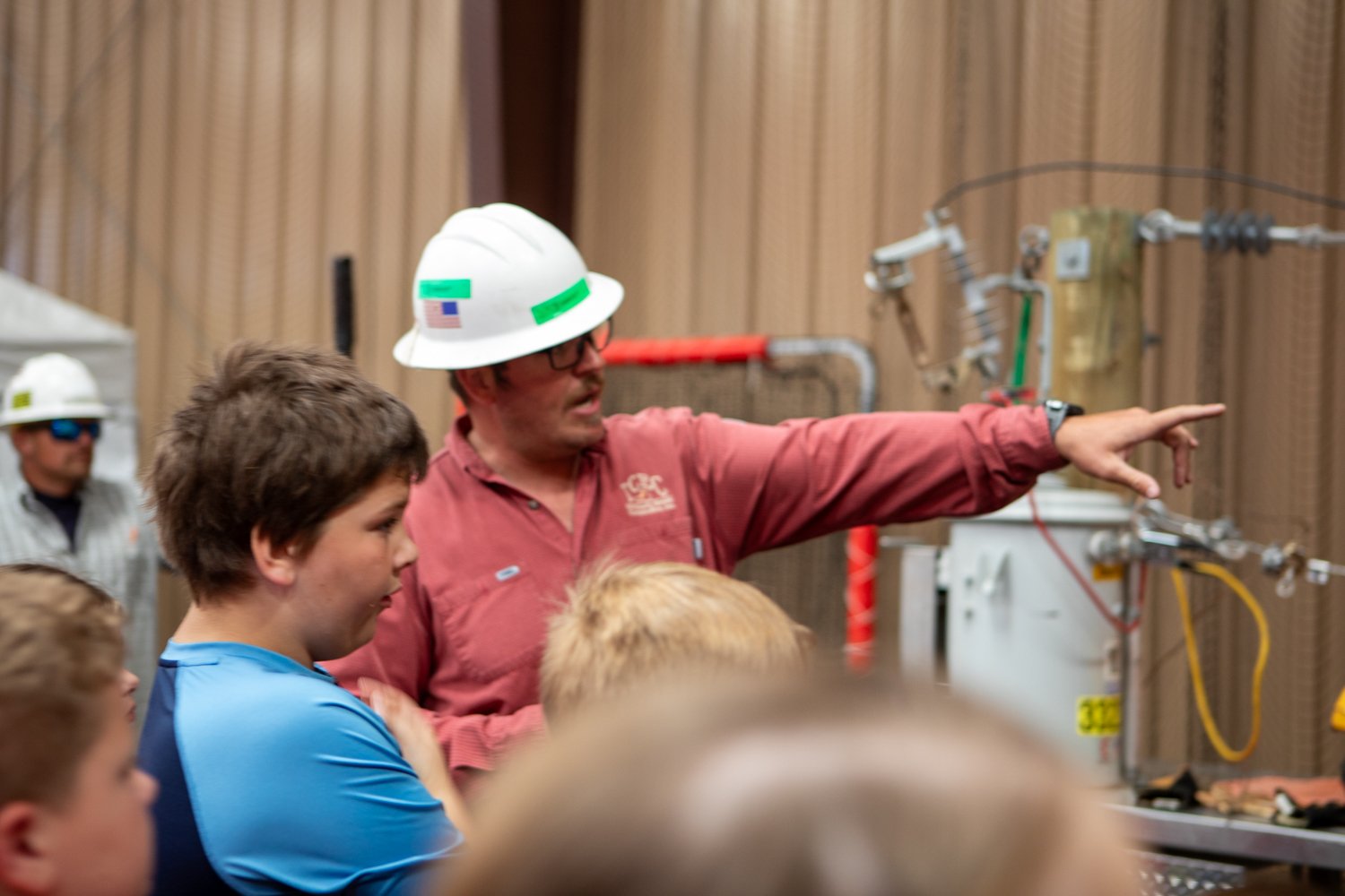A man wearing a hard hat demonstrates equipment to a group of grade school students, pointing as they watch and listen during an interactive learning activity.