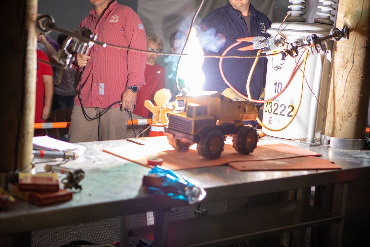 Two men in work shirts are performing a science demonstration on a metal table for a group of children. A bright spark and a small flame are visible coming from a yellow toy truck. There is a white electrical transformer nearby with wires and clamps attached to it.