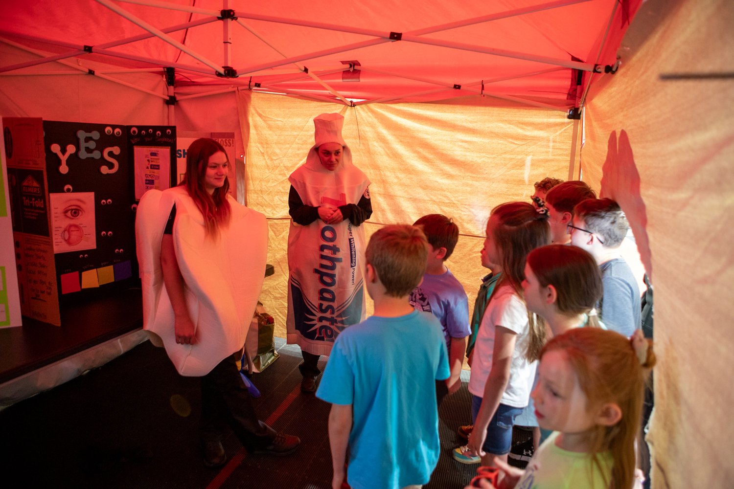Inside a red-topped tent, two women dressed in dental-themed costumes—one as a large white molar tooth and the other as a tube of toothpaste—interact with a group of elementary-aged children. To the left, a trifold display board titled "YES" features educational diagrams of an eye and various colorful posters. The children are gathered around, listening to the presentation on dental hygiene.