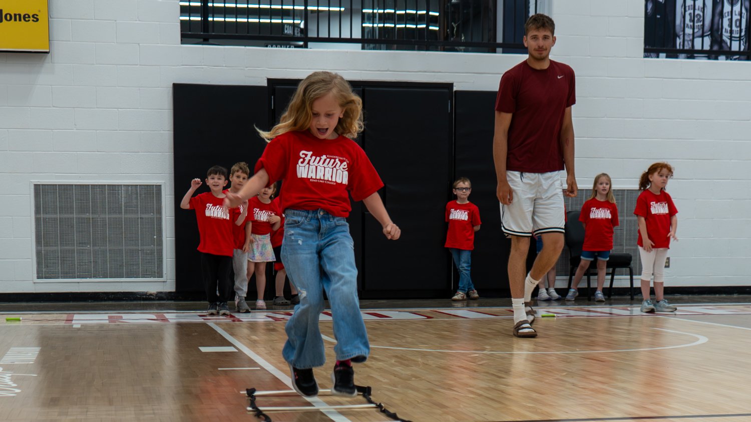 A young girl in a red "Future Warrior" shirt and jeans jumps through an agility ladder on a gymnasium floor. Other children in matching shirts and a tall instructor in athletic gear watch her progress during the 2026 Kindergarten Day event.