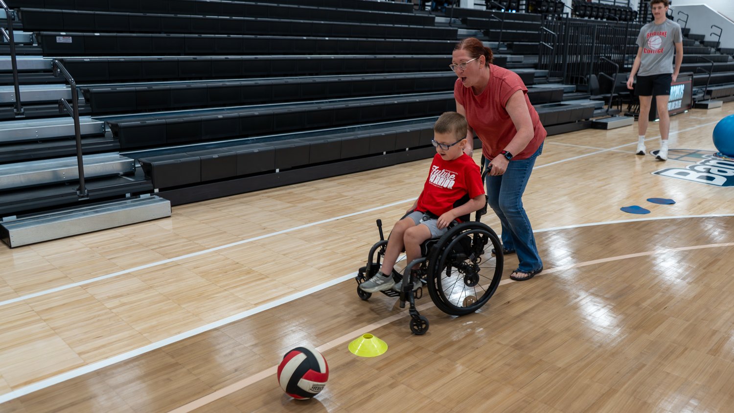 A woman assists a young boy in a wheelchair during an athletic activity on a gymnasium floor. The boy, wearing a red "Future Warrior" shirt, maneuvers toward a volleyball and a yellow cone as they participate in the event.