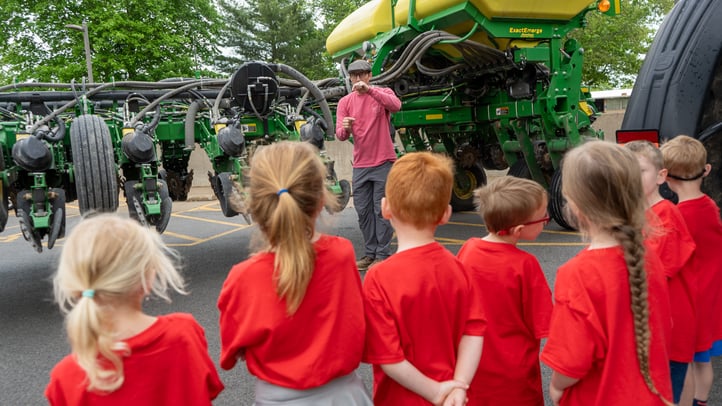 A man in a pink shirt and cap stands before a large green agricultural planter, gesturing as he teaches a group of children. The kids, all wearing matching red T-shirts, listen to the presentation outdoors.
