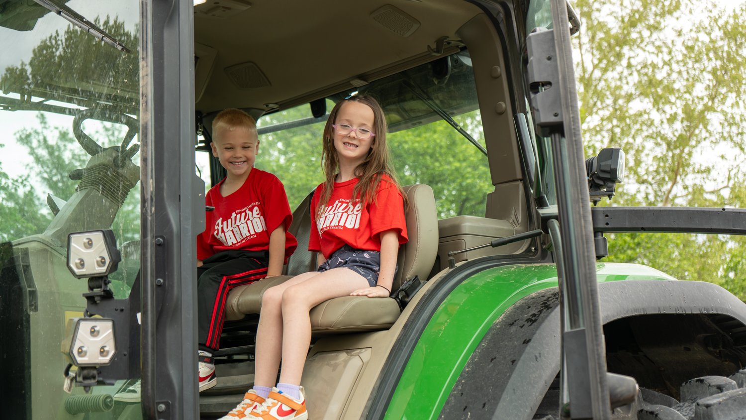Two children wearing red "Future Warrior" shirts sit inside the cab of a large green tractor, smiling. They are exploring the heavy equipment during the Rend Lake College Kindergarten Day event.