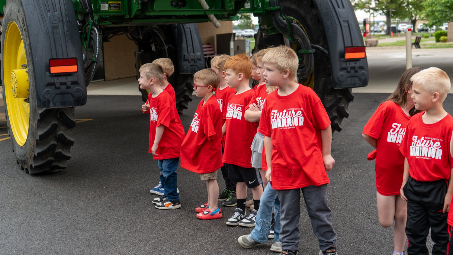 A group of kindergarteners in matching red "Future Warrior" shirts stand in a line, looking up at a massive piece of green agricultural machinery. The children appear curious as they observe the scale of the heavy equipment during Rend Lake College's Kindergarten Day.