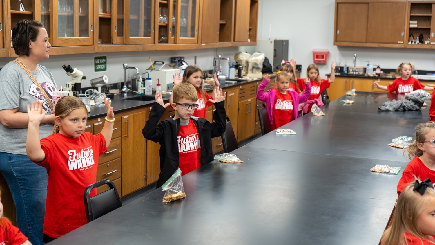 In a science lab, children in red "Future Warrior" shirts stand at long tables with their hands raised. An adult looks on as the students participate in an activity featuring lab equipment and snacks.
