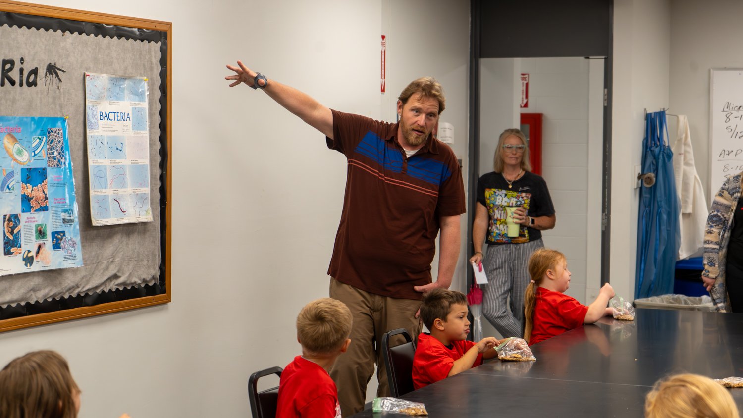  In a science classroom, an instructor gestures toward a bulletin board labeled "Bacteria" while teaching a group of kindergarteners. The children, wearing red "Future Warrior" shirts, sit at a long table with snacks as they listen to the presentation.