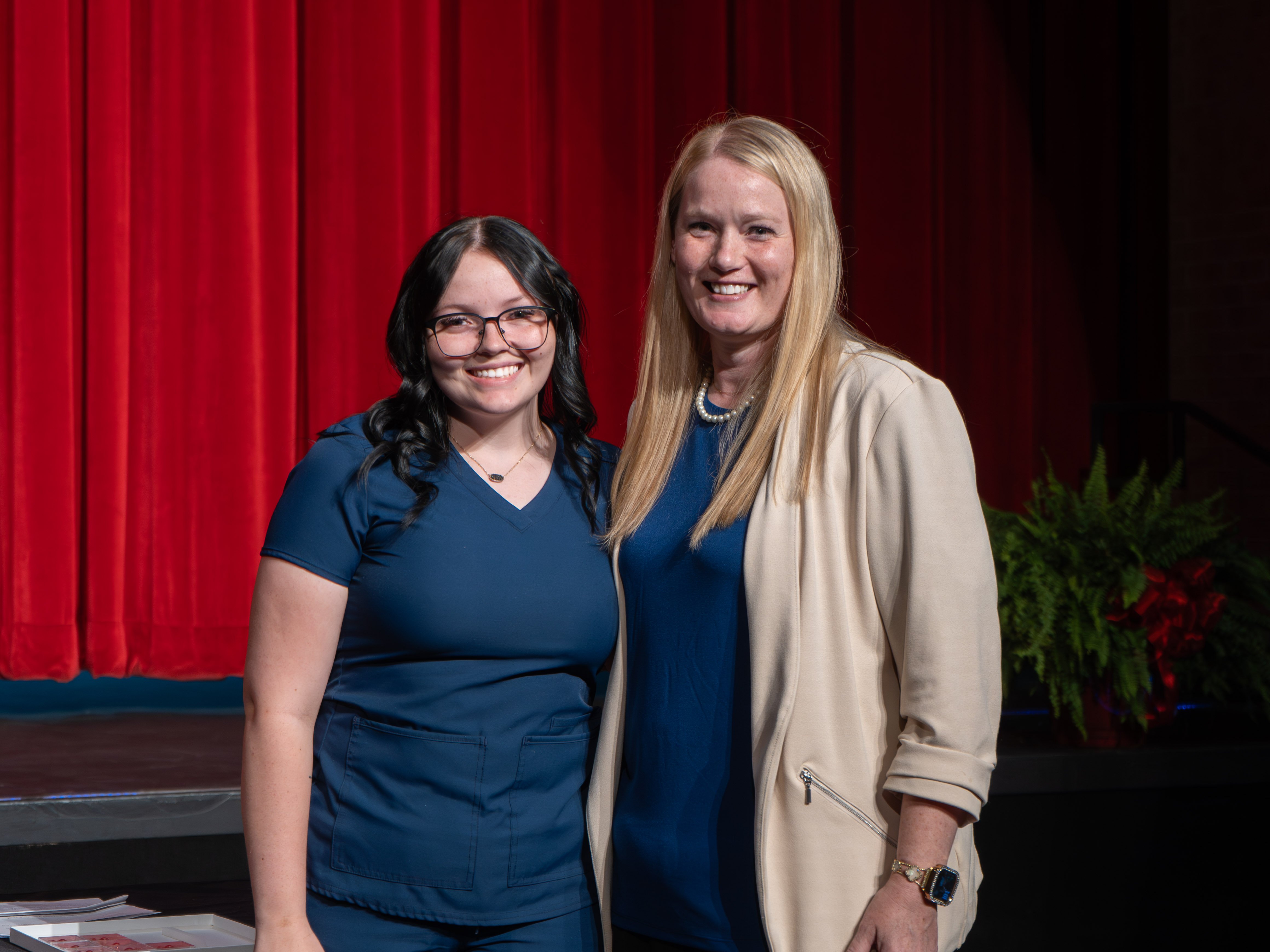 Medical assistant graduate Londynn Browning, wearing glasses and navy blue scrubs, smiles next to her instructor on a stage. The instructor is a blonde woman wearing a cream blazer and blue top. A large red curtain serves as the background.