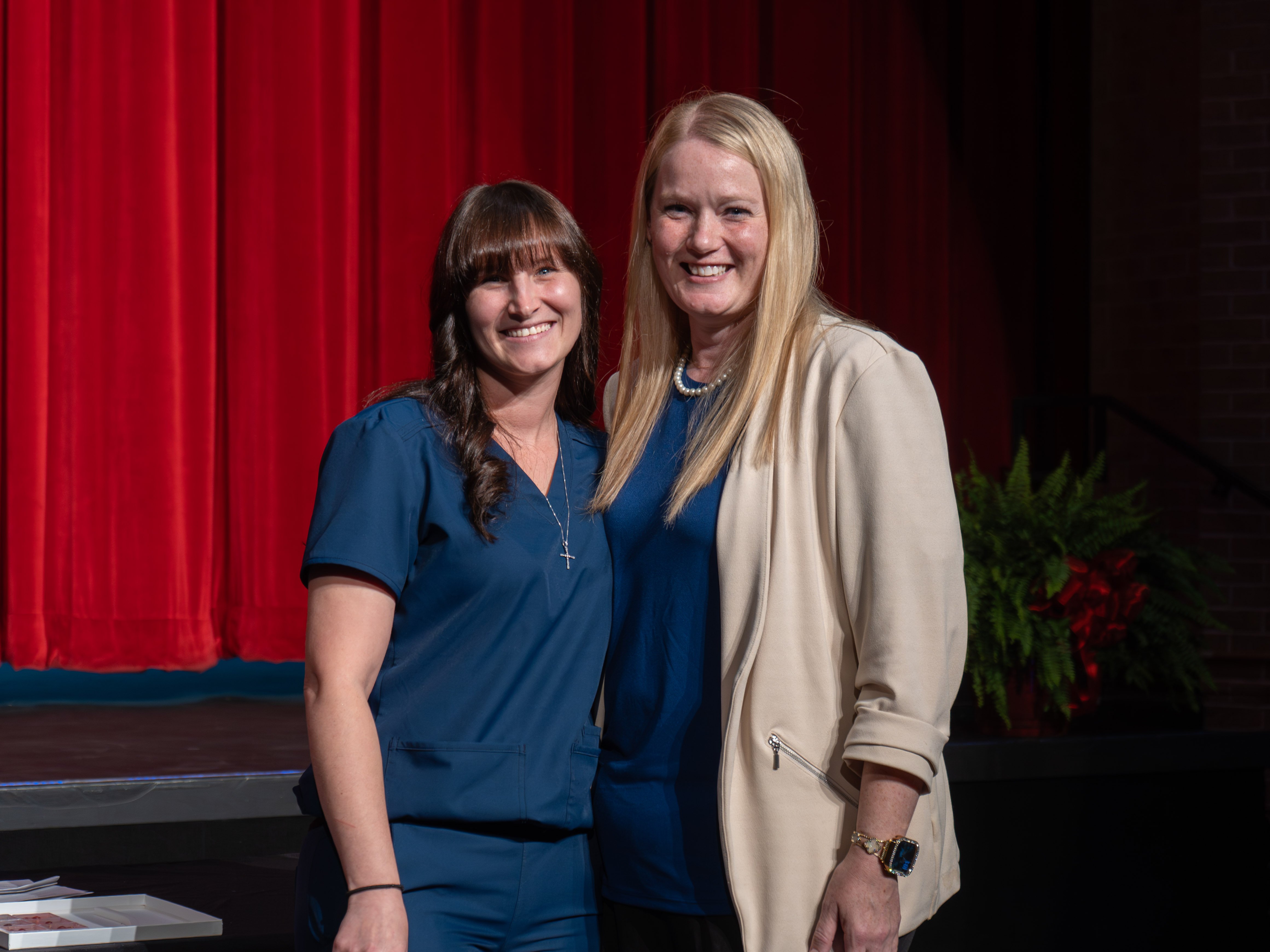 Medical assistant graduate Jessi Byerly smiles while wearing navy blue scrubs and a silver cross necklace. She stands next to her blonde instructor, who is wearing a cream blazer, on a stage in front of a vibrant red curtain.