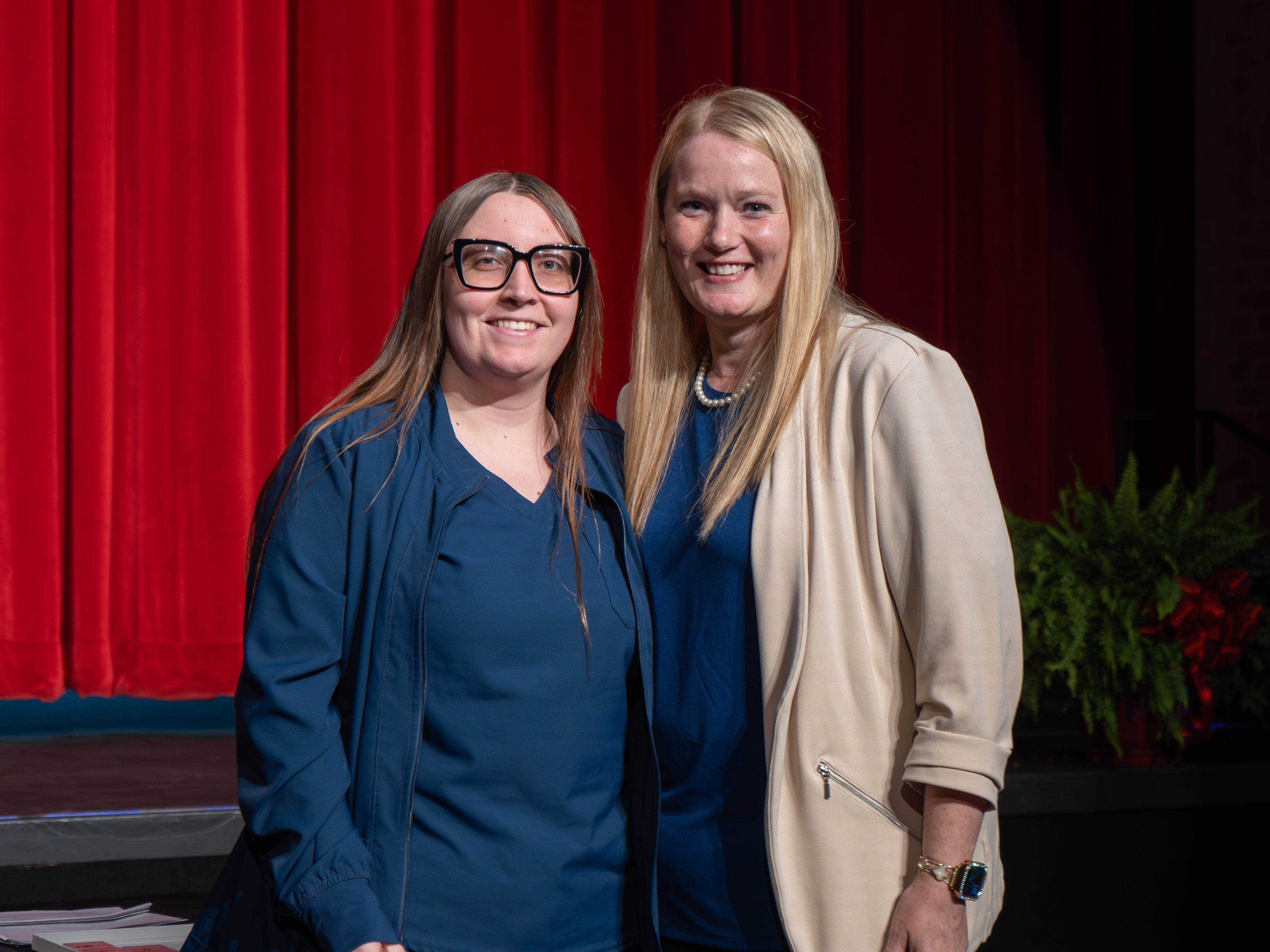 Medical assistant graduate Anna Ellis smiles while wearing thick black-rimmed glasses and a navy blue scrub jacket. She stands on a stage next to her instructor, a blonde woman wearing a cream blazer. The background features a large red curtain.