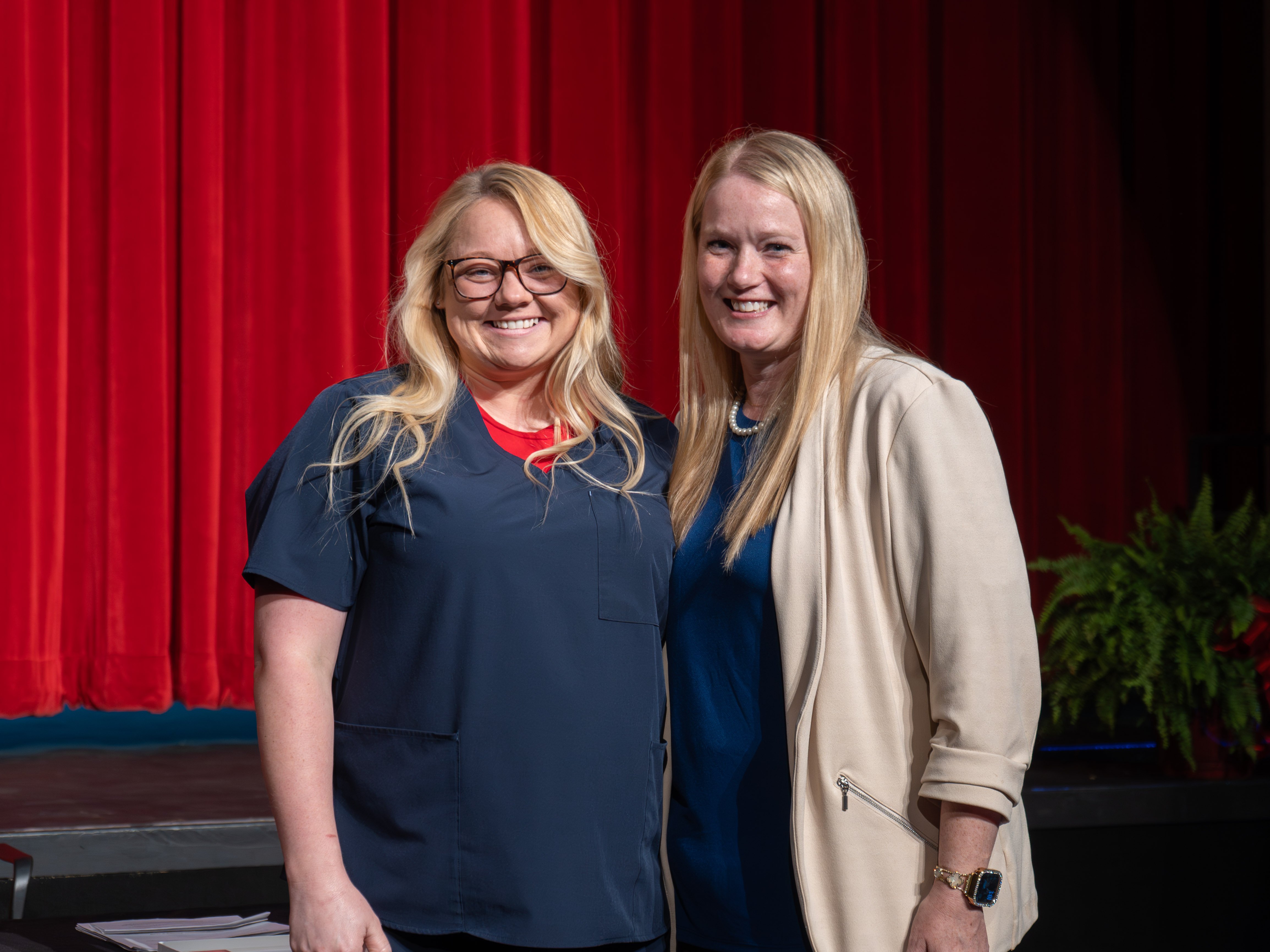 Medical assistant graduate Blaise Emling, wearing glasses and navy blue scrubs with a red undershirt, smiles alongside her instructor. The instructor stands to her right in a cream blazer and blue top. They are posing on a stage in front of a red curtain.