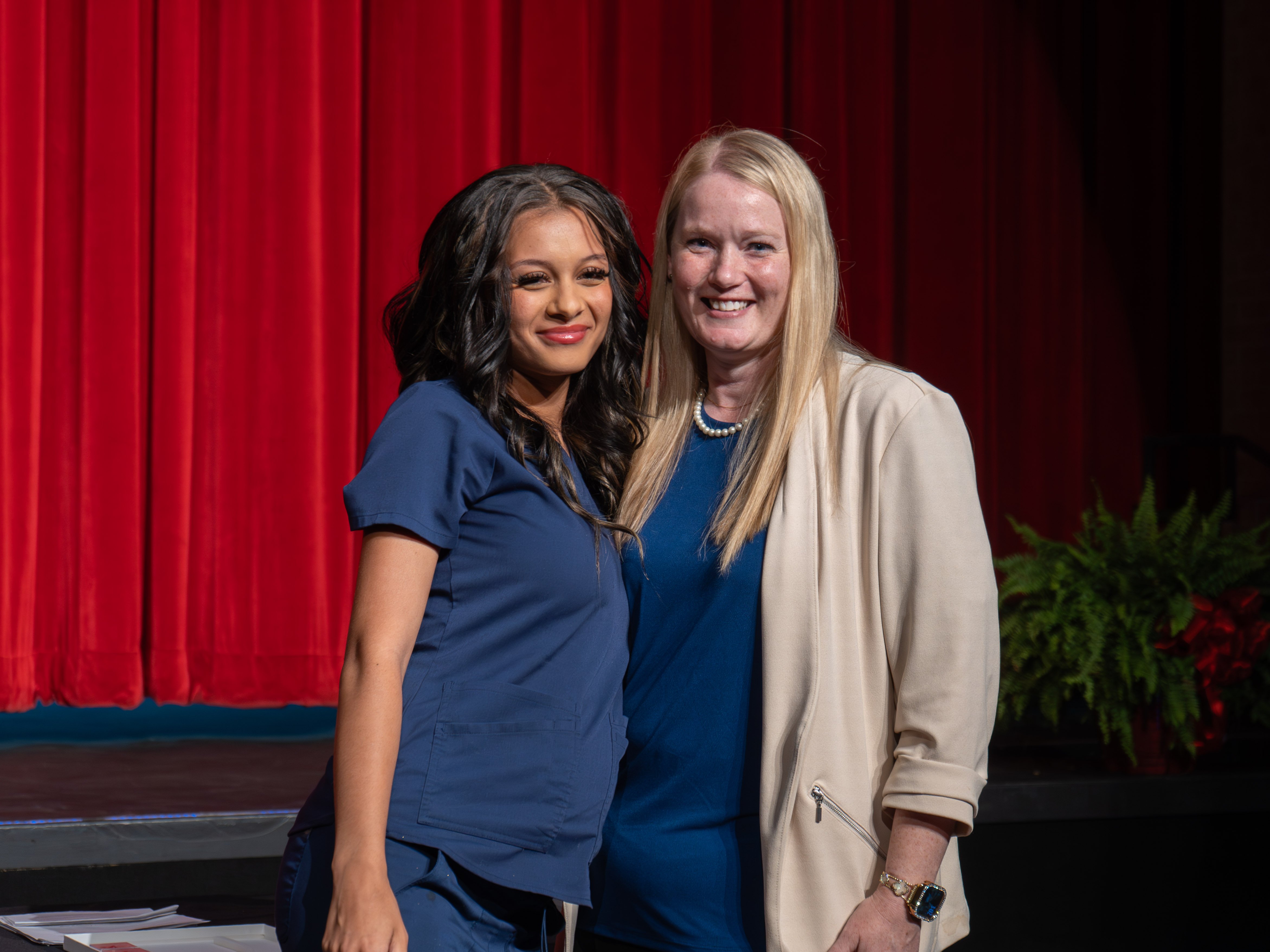 Medical assistant graduate Kalia Grundy poses with her instructor on a stage in front of a red curtain. Kalia has long, dark wavy hair and is wearing navy blue scrubs. The instructor, a smiling blonde woman in a cream blazer and blue top, stands to her right with an arm around her.