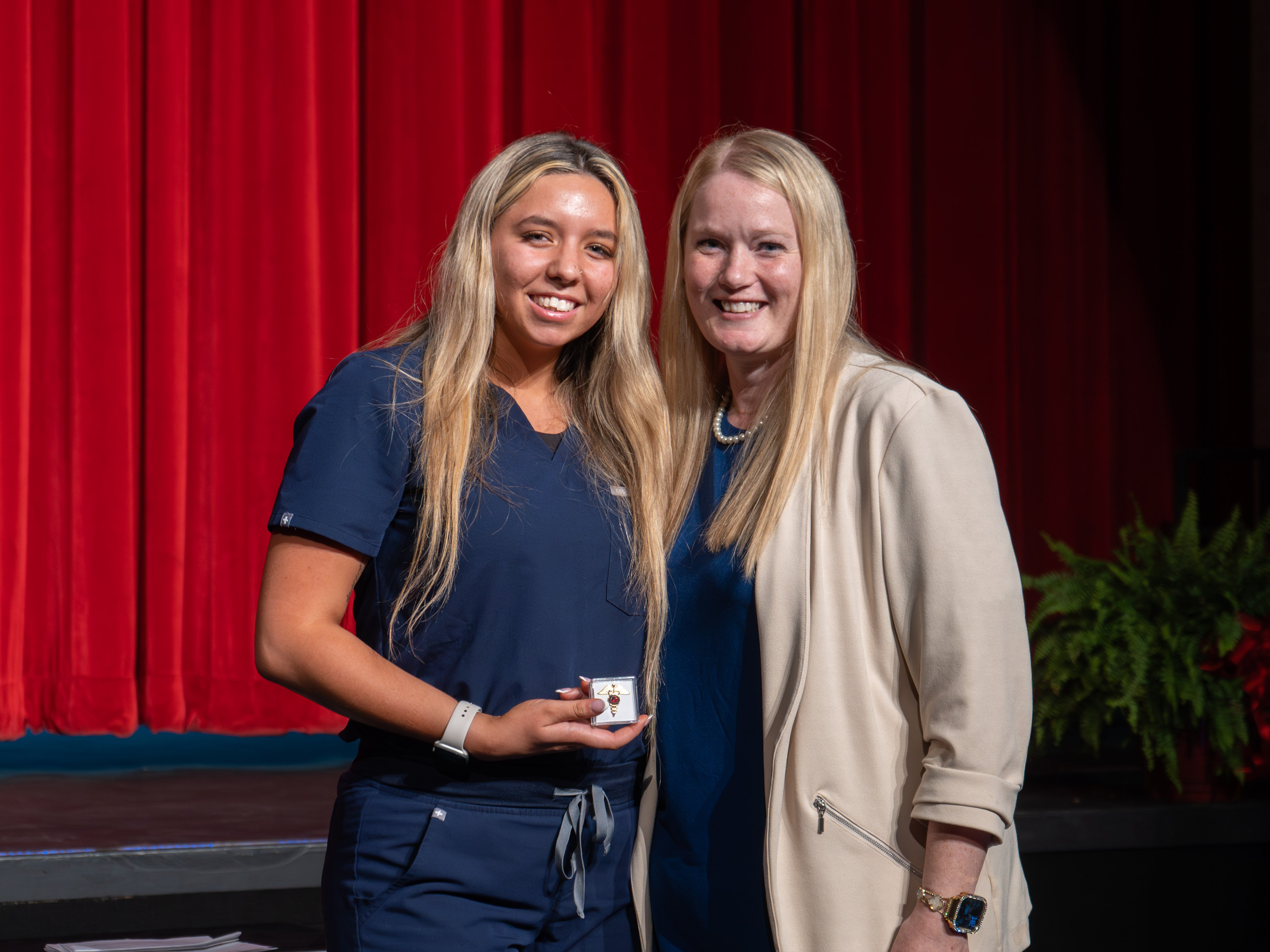 Medical assistant graduate Erin Harpstrite smiles while holding a small, clear box containing a graduation pin. She has long blonde hair and wears navy blue scrubs. She is standing next to her instructor, a blonde woman in a cream blazer, on a stage with a red curtain background.