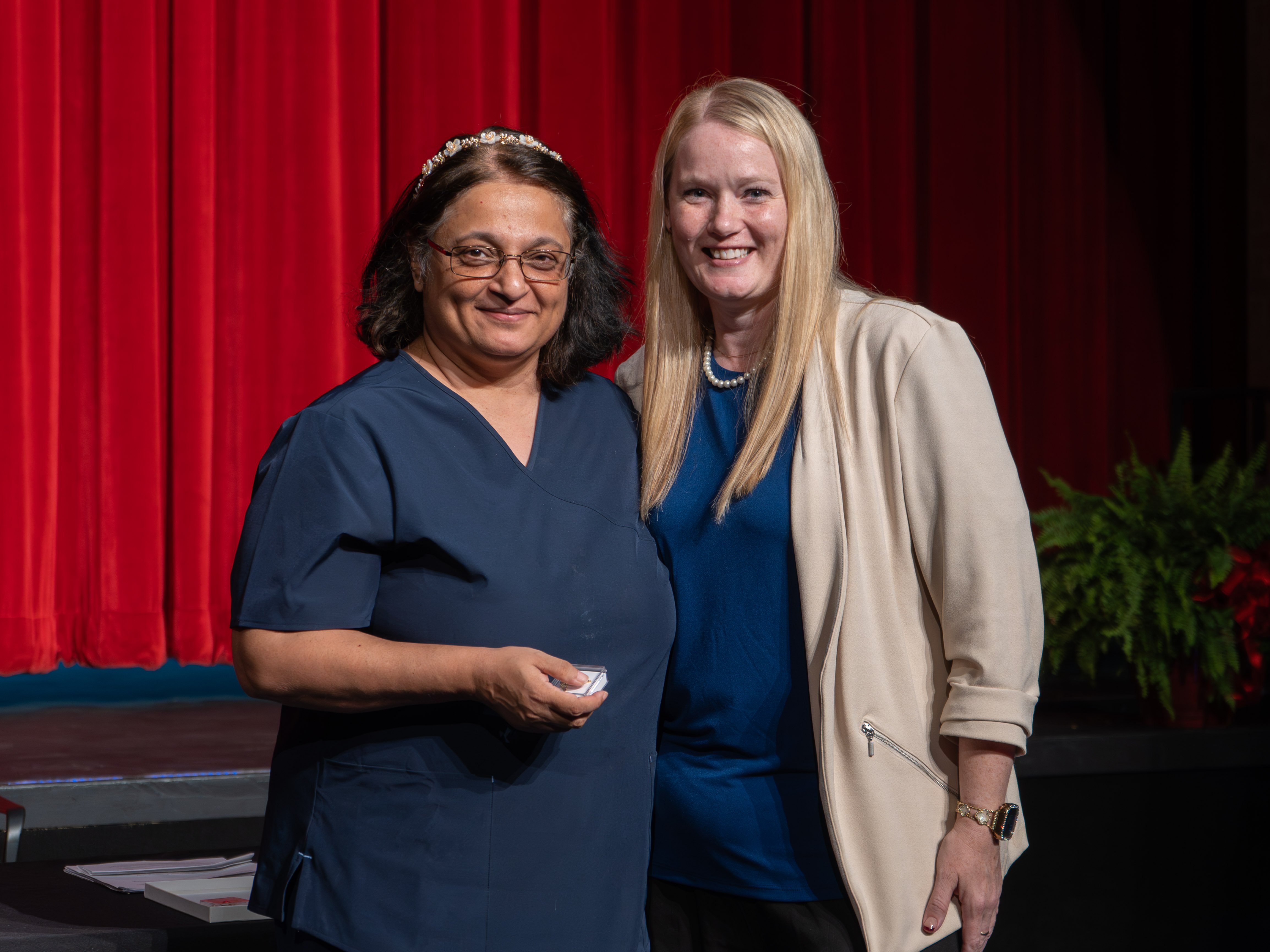 Medical assistant graduate Priya Nair, wearing glasses, a beaded headband, and navy blue scrubs, smiles while holding her graduation pin box. She is posing on a stage next to her instructor, a blonde woman wearing a cream blazer and a pearl necklace, in front of a red curtain.