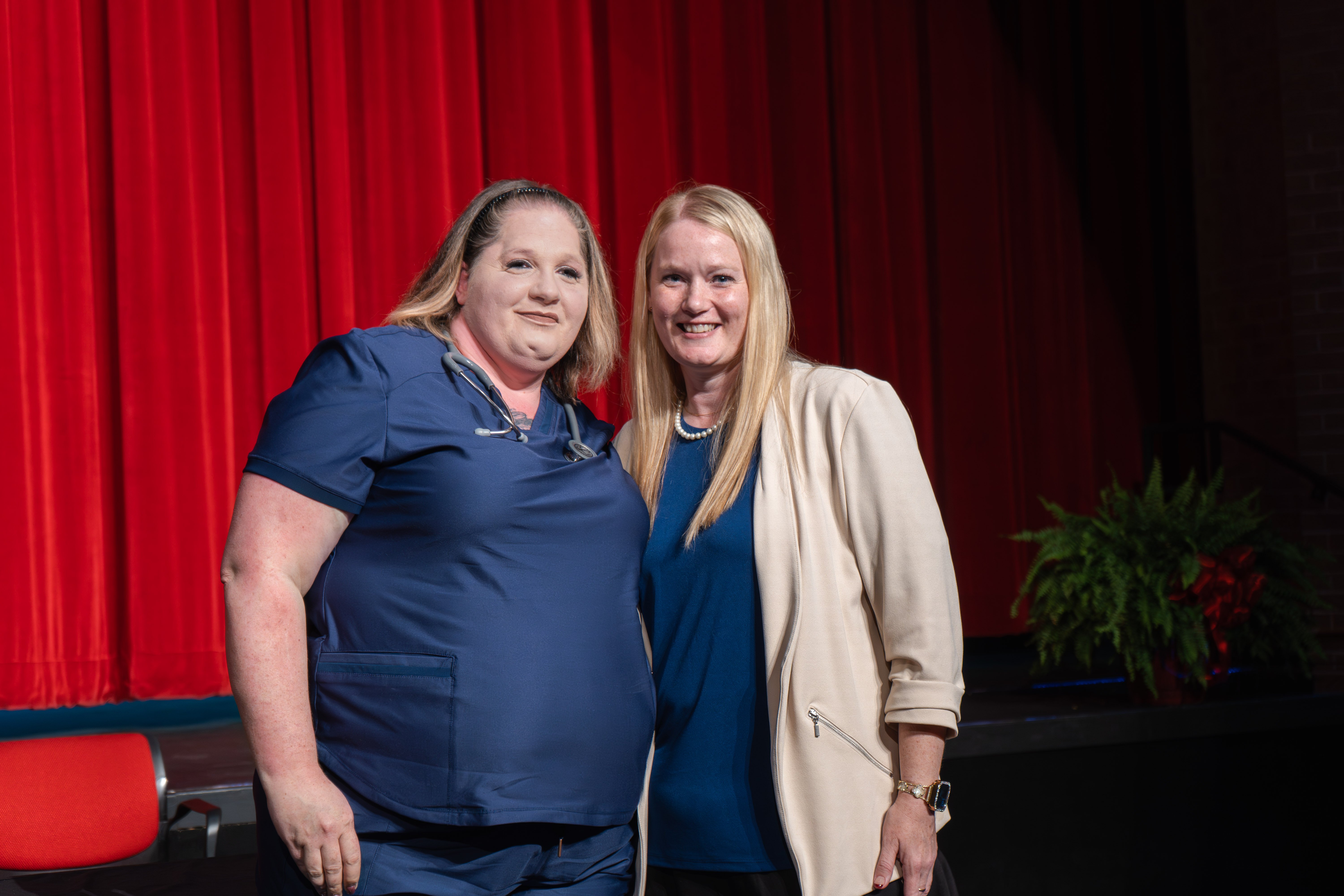 Medical assistant graduate Michelle Arriaga smiles while posing with her instructor on a stage with a red curtain backdrop. Michelle wears navy blue scrubs with a stethoscope around her neck. The instructor stands next to her wearing a cream blazer over a blue shirt.