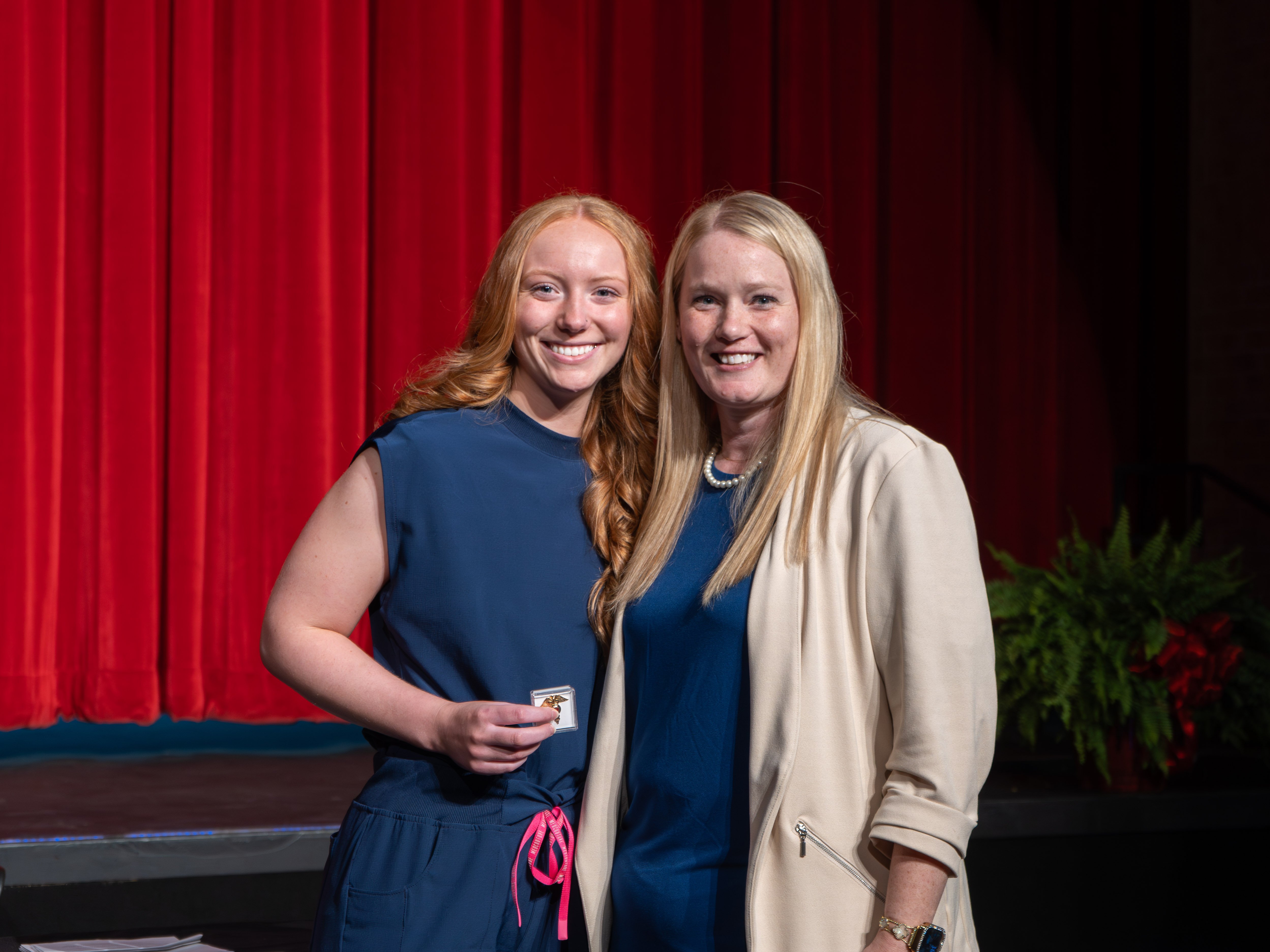 Medical assistant graduate Ryleigh Bates smiles broadly while holding a small box with her graduation pin. She has long red hair and is wearing a sleeveless navy blue scrub top with a pink drawstring. She is standing next to her smiling instructor, who wears a cream blazer, in front of a red curtain stage.
