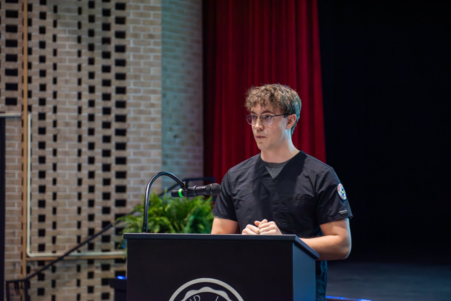 A close-up shot of nursing student Michael Parks standing at a dark podium with a microphone, delivering a speech on a stage. He is wearing glasses and black medical scrubs with an institutional patch on the left sleeve. A brick wall and a red stage curtain are visible in the background.